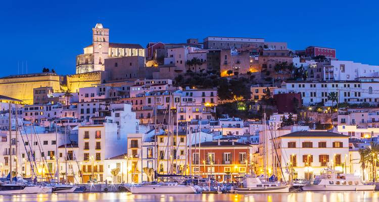 Avondgezicht van Ibiza-stad met een haven vol jachten en verlichte gebouwen.