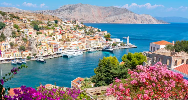 Vue panoramique surélevée de la ville et du port colorés de Symi, encadrés par des fleurs roses et la mer bleue