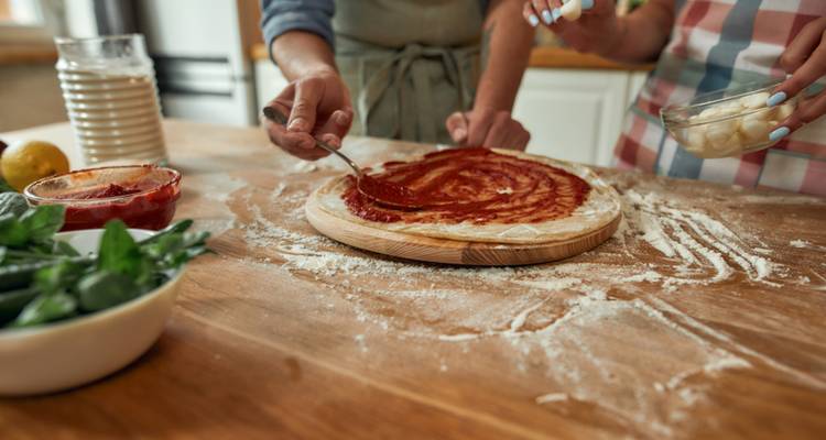 Two people preparing pizza dough with tomato sauce on a wooden table.