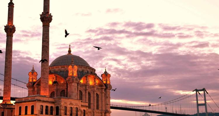 Golden beleuchtete Ortaköy-Moschee vor einem dramatischen pastellfarbenen Sonnenuntergangshimmel mit der Bosporus-Brücke als Silhouette.