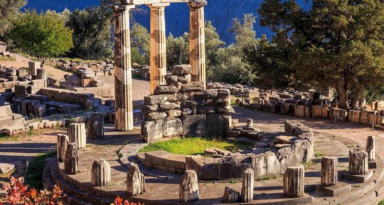 Les ruines circulaires de la Tholos de Delphes avec trois colonnes debout entourées de cyprès et un arrière-plan montagneux.