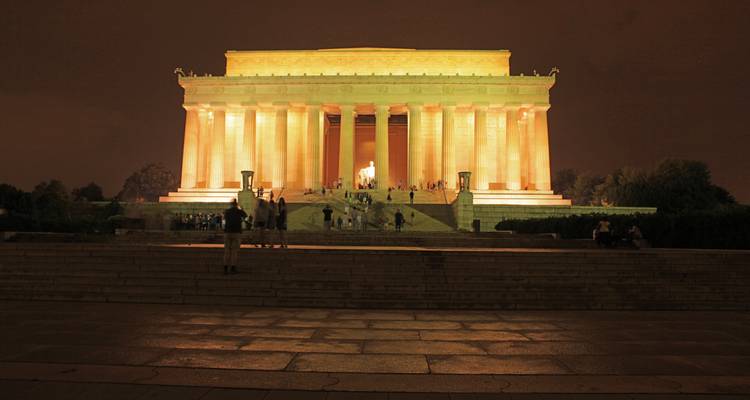 Le Lincoln Memorial illuminé brille d'une teinte dorée contre le ciel nocturne tandis que de petits visiteurs s'attardent sur les marches.