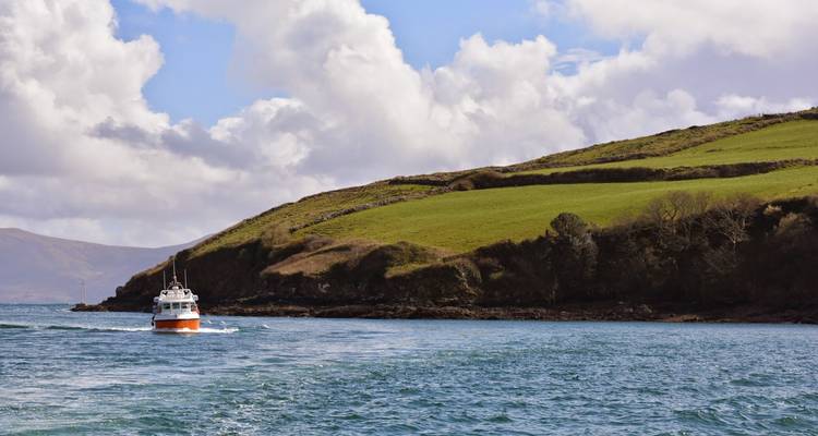 A small passenger boat sails past green rolling hills into a calm blue bay under partly cloudy skies.
