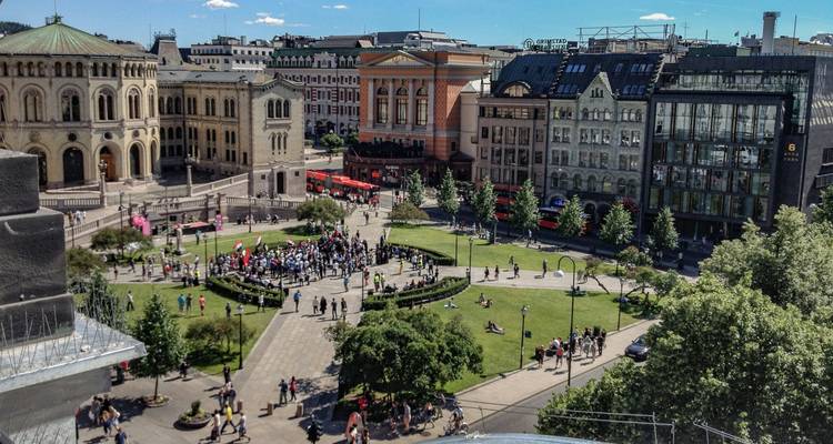 Eine Luftaufnahme eines belebten Stadtplatzes mit historischen Gebäuden, grünen Rasenflächen und Menschenmengen, die sich unter einem strahlend blauen Himmel versammeln.