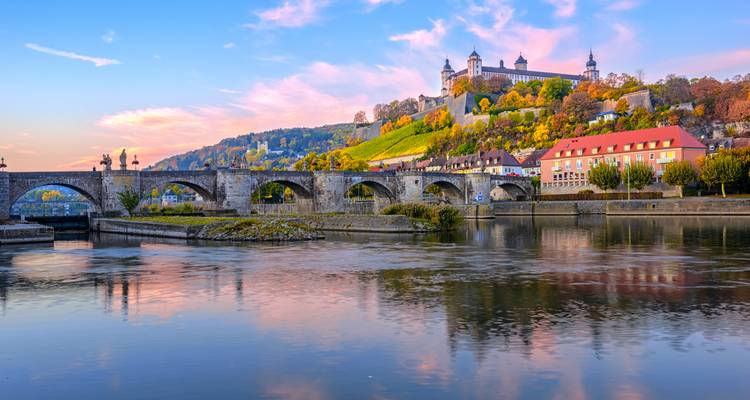 A historic bridge and fortress with a reflection on a river during sunset.