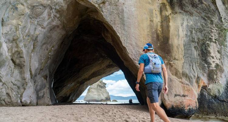 Rucksacktourist betritt den natürlichen Felsbogen der Cathedral Cove, der sich zu einem Sandstrand und einer Felssäule hin öffnet.