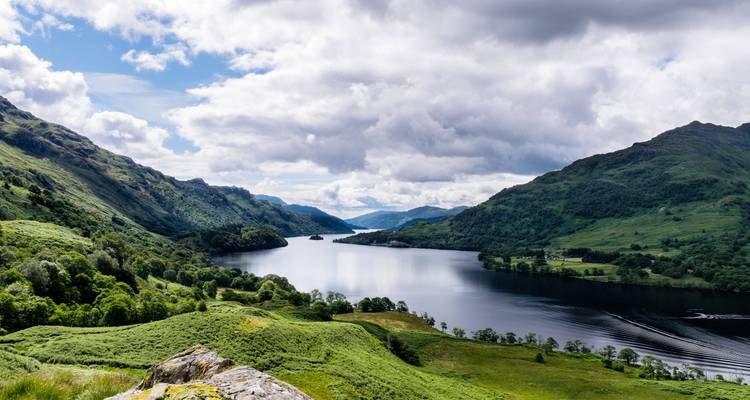 Vue panoramique sur un loch écossais tranquille, encadré par des montagnes vertes ondulantes sous un ciel dramatique.