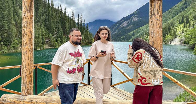Three people standing on a wooden deck by a mountain lake.