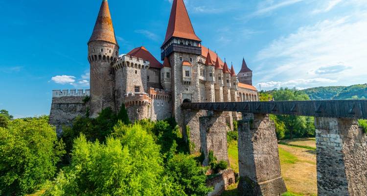 Le château de Corvin, digne d'un conte de fées, avec ses toits rouges et ses tours de pierre, s'élève au-dessus d'une végétation luxuriante et d'un pont en bois.