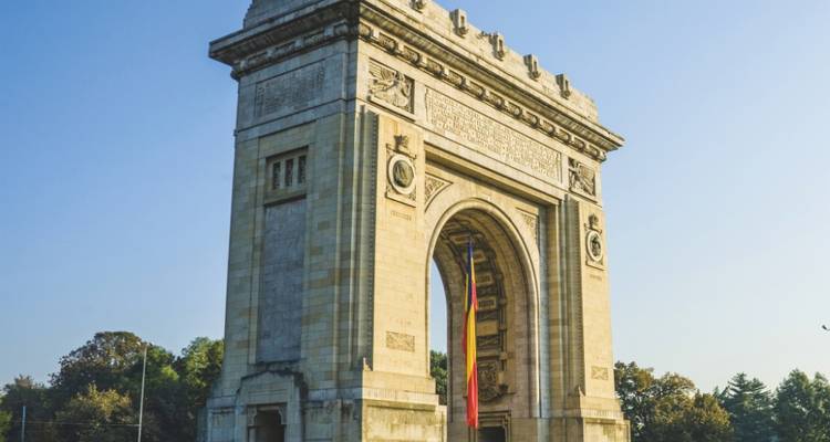 Le monument en pierre Arcul de Triumf se dresse majestueusement contre un ciel matinal dégagé, entouré d'arbres.