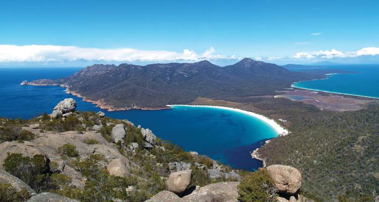Vue panoramique d'une baie aux sables blancs et courbes, encadrée par des pics de granit escarpés et une mer turquoise.