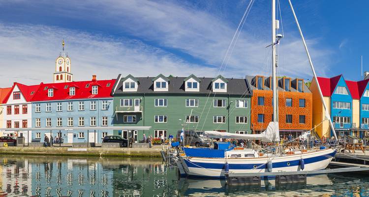 Kleurrijke Noordse huizen aan het water en een aangemeerde zeilboot weerspiegeld in kalm havenwater onder een helder blauwe hemel.