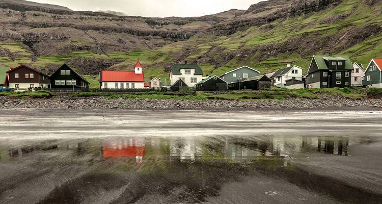Afgelegen Faeröers dorp met een wit-en-rood kerkje en met gras bedekte rotsen weerspiegeld op een zwart zandstrand bij laag water.