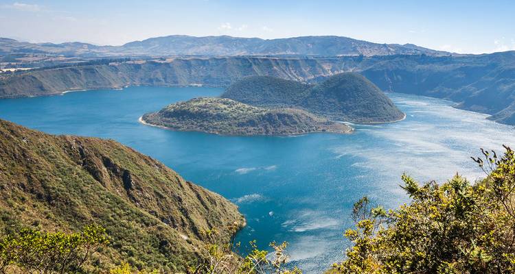 Lago de cráter azul profundo con islotes exuberantes rodeado de crestas andinas y cielos despejados.
