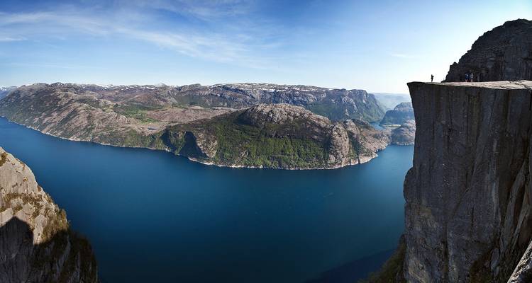 Saliente rocoso escarpado con vista a un fiordo azul profundo y montañas, con figuras diminutas en la cima