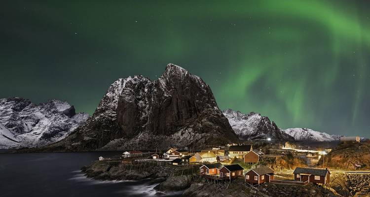 Las auroras boreales bailando sobre cabañas de pesca cubiertas de nieve y una montaña de granito macizo en una noche de invierno.