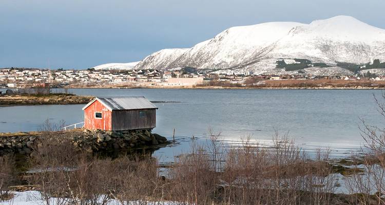 Una solitaria casita de botes roja en un peñasco frente a un pueblito costero nevado y montañas con cimas blancas.