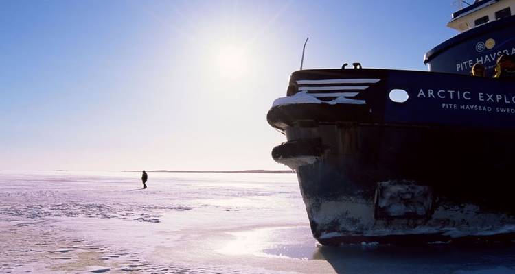 El gran rompehielos Explorador Ártico descansa en el mar congelado mientras una figura solitaria camina sobre el hielo bajo un sol brillante.