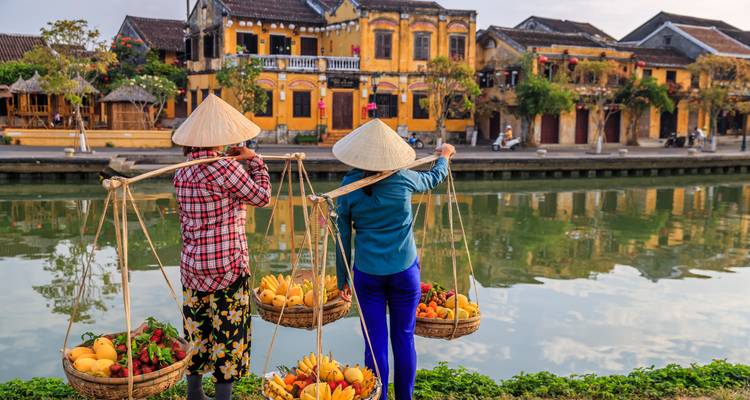 Two women in traditional Vietnamese attire carrying fruit baskets beside a canal, with old buildings in the background.