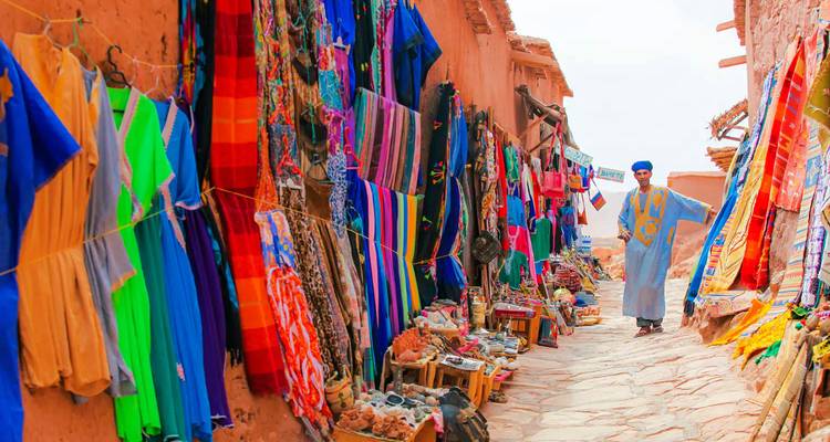 Earthen alley lined with vibrant textiles and handicrafts where vendors and tourists browse.