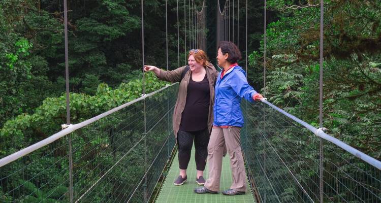 Two women pause on a suspended canopy bridge high above lush green rainforest.
