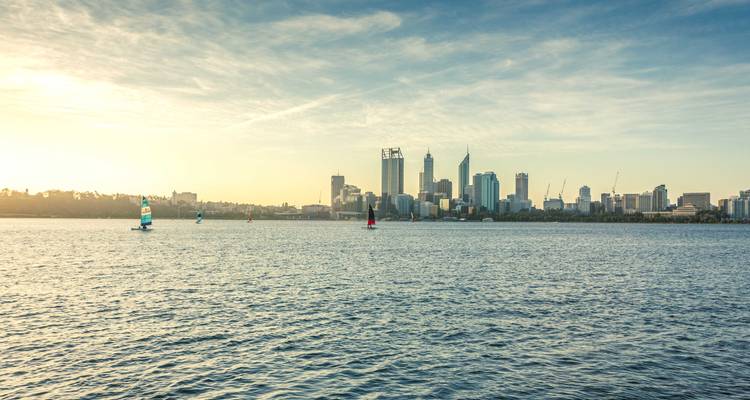 Die Silhouette der Skyline von Perth bei Sonnenaufgang, die sich in den ruhigen Gewässern des Swan River spiegelt, mit kleinen Segelbooten in der Ferne.