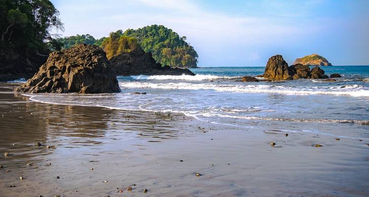 Schilderachtig strand met rotspartijen, zachte golven en eilanden op de achtergrond.