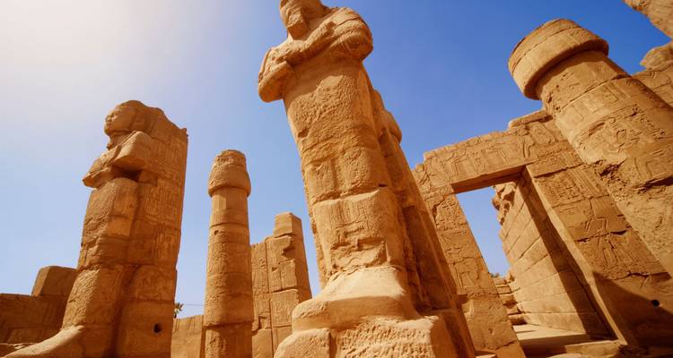 Vista hacia arriba de las enormes estatuas de piedra y columnas del Templo de Karnak contra un cielo azul despejado.