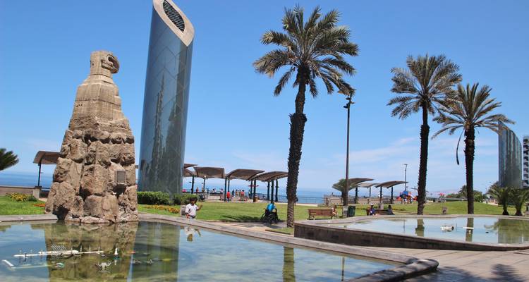 Parque de esculturas modernas junto al mar con gente caminando y palmeras.