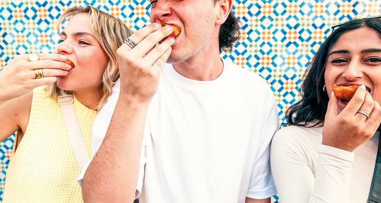 Three friends bite into pastel de nata pastries against a colorful Portuguese tile wall.