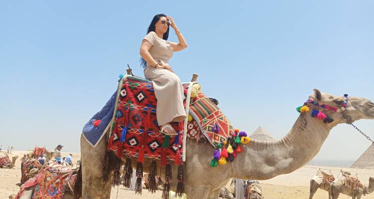 Woman riding a camel with the pyramids visible in the distance.