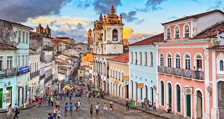 Kleurrijke koloniale gebouwen staan langs een geplaveide straat vol voetgangers, met een sierlijke barokke kerk die erboven uittorent onder een warme late-middaghemel.