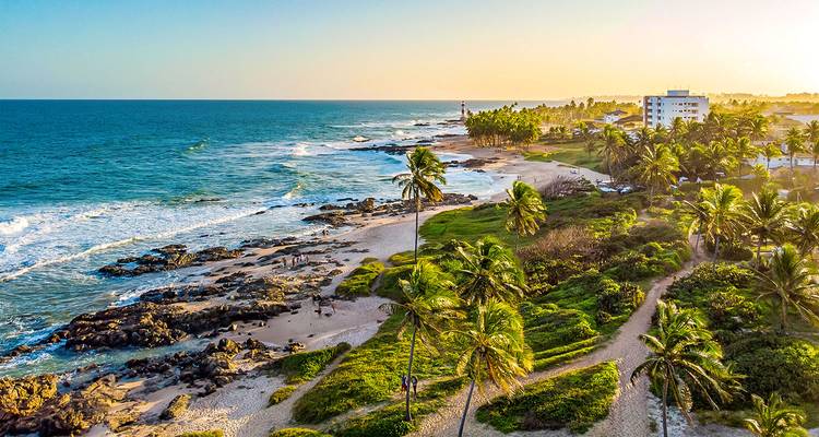 Luchtfoto van de kust met een door palmbomen omzoomd zandstrand met rotsformaties en turquoise oceaangolven bij zonsondergang, een vuurtoren en resortgebouwen in de verte.
