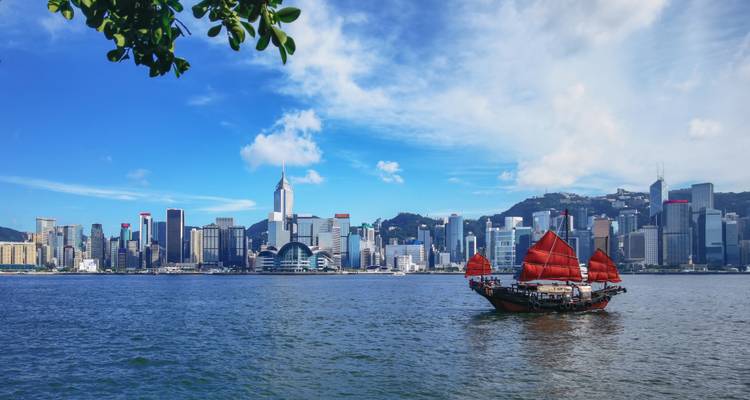 Ein traditionelles Dschunkenboot mit roten Segeln überquert den Victoria Harbour mit Hongkongs moderner Skyline im Hintergrund.