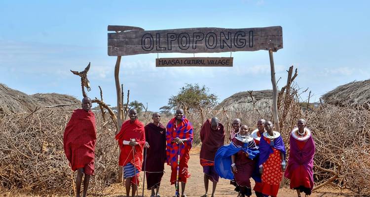 Groupe de Maasaï marchant sous le panneau d'entrée du village culturel d'Olpopongi entouré d'une clôture en broussailles épineuses.