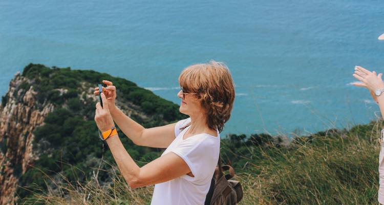 Una viajera fotografía un acantilado costero dramático y un mar azul desde un mirador con césped.