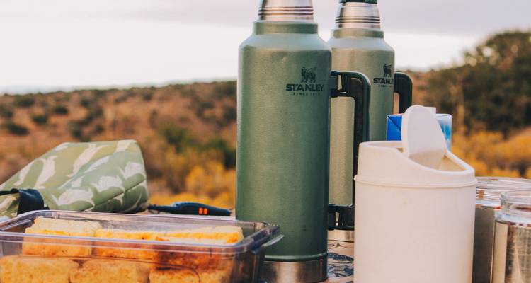 Configuración de picnic al aire libre con termos, snacks y tazas sobre una mesa en tierra semi-árida con matorrales.