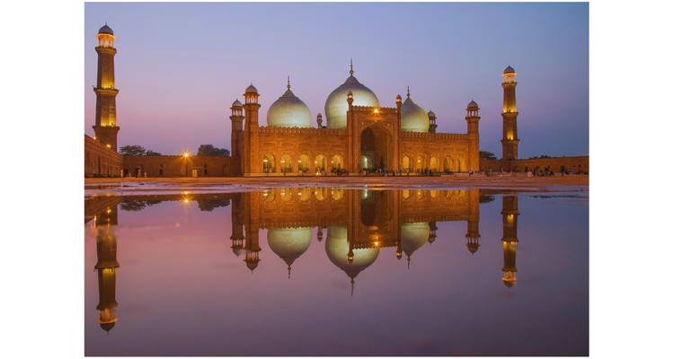 Illuminated Badshahi Mosque reflected in a shallow pool at dusk with pink and purple sky.