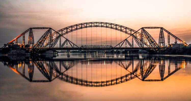Steel railway bridge spanning a wide river perfectly mirrored in calm water at sunset.