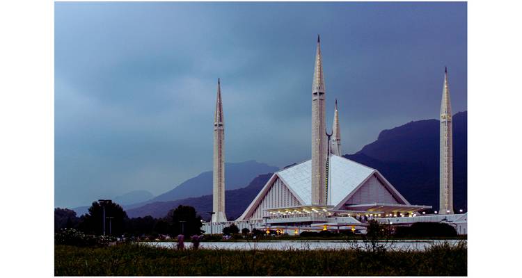 Modern white Faisal Mosque with dramatic minarets set against moody mountains and twilight sky.