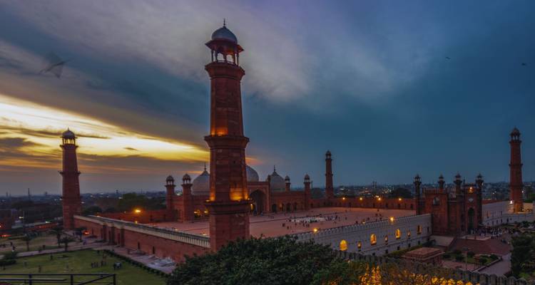 Evening panorama of Badshahi Mosque glowing under dramatic sunset clouds with visitors in the courtyard.