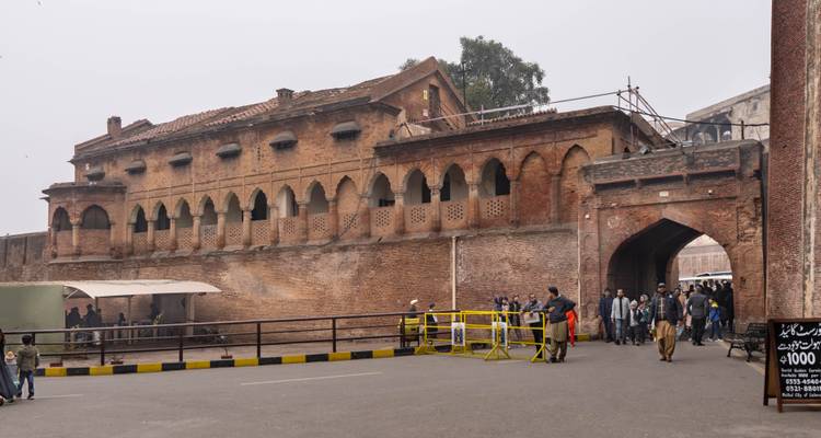 Side view of historic red-brick barracks within Lahore Fort complex with tourists walking near security barriers.