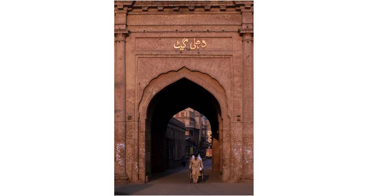 Tall brick Mughal gateway with Urdu inscription and two pedestrians walking through the arch at dusk.