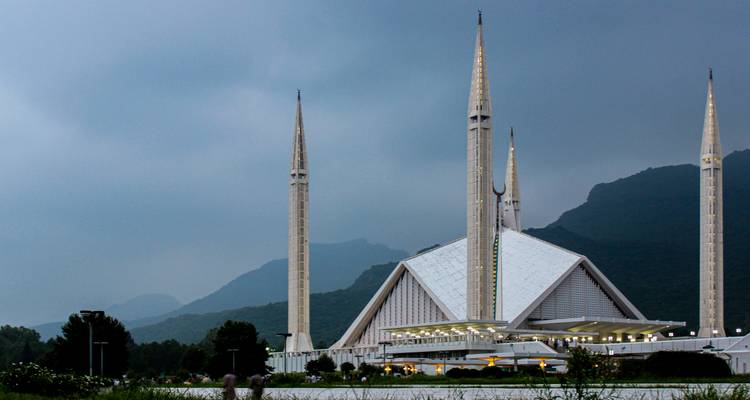 Striking modern Faisal Mosque framed by lush greenery with misty hills and moody grey sky behind.