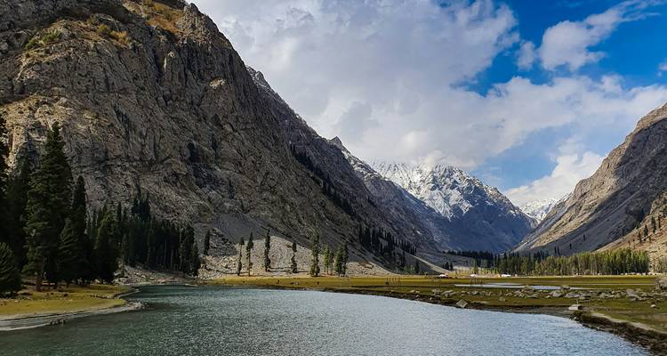 A tranquil river winds through a dramatic mountain valley framed by steep rocky cliffs and distant snow-covered peaks beneath a vivid blue sky.