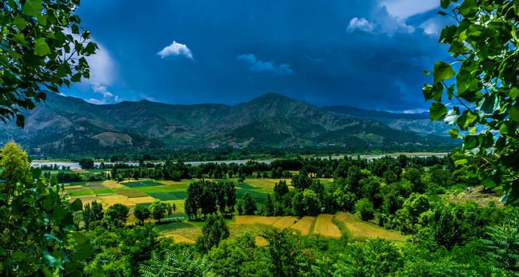 Lush green fields, forests and a wide valley stretch toward rolling mountains under a moody, cloud-filled sky.