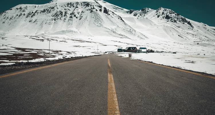 A straight empty road leads toward towering snow-covered mountains under a brilliant turquoise sky, evoking a sense of adventure.