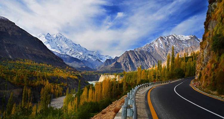 A curving highway skirts golden autumn trees with dramatic snow-capped peaks and a deep valley river in the distance.