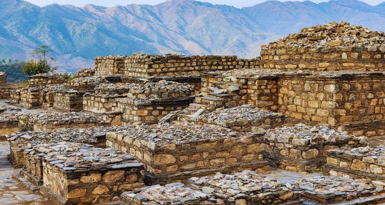Des ruines de pierre éparses d'une ancienne cité bouddhiste se dressent sous des montagnes escarpées et un ciel aux teintes bleues.