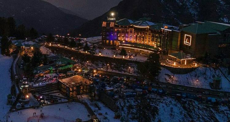 Vue aérienne nocturne d'une station de montagne enneigée illuminée de lumières colorées avec une patinoire en contrebas.